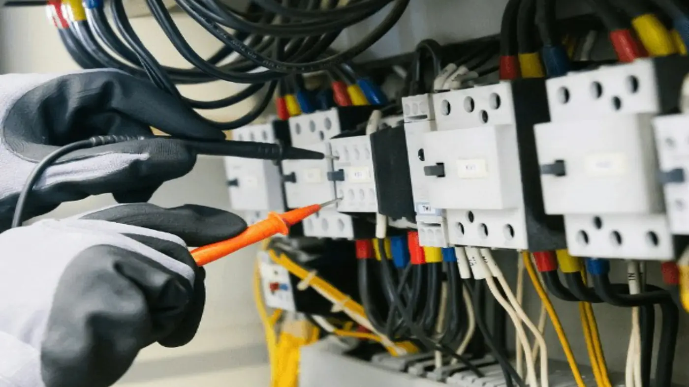 A gloved hand testing wiring inside an open electrical panel with a meter probe