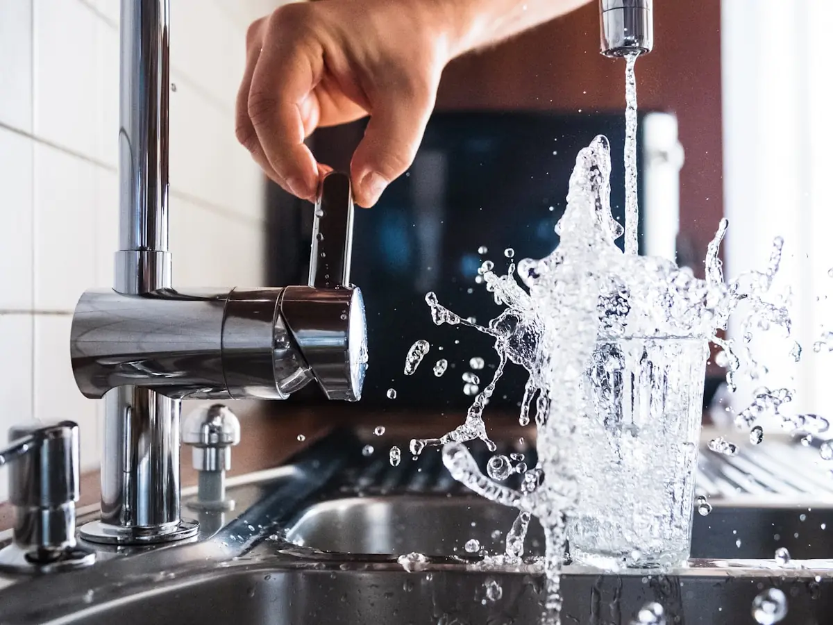 A hand opening a kitchen tap with water splashing into a stainless steel sink