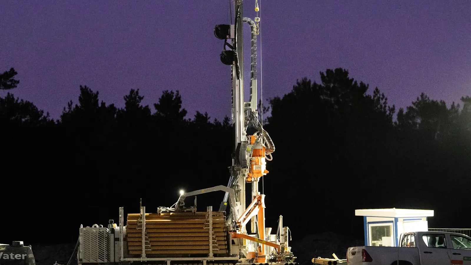 A water well drilling rig lit up and working at dusk beside a stand of pines