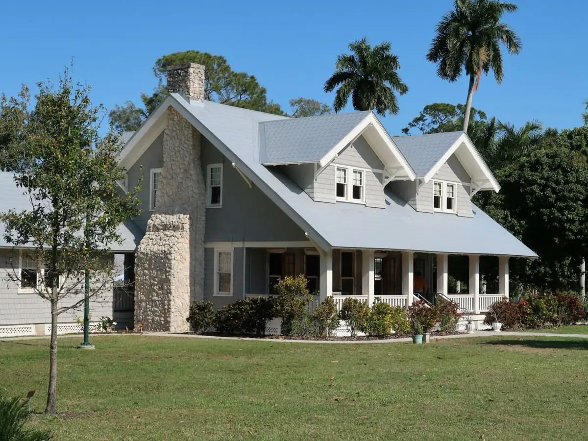 A gray two-story house with a stone chimney, porch and palm trees