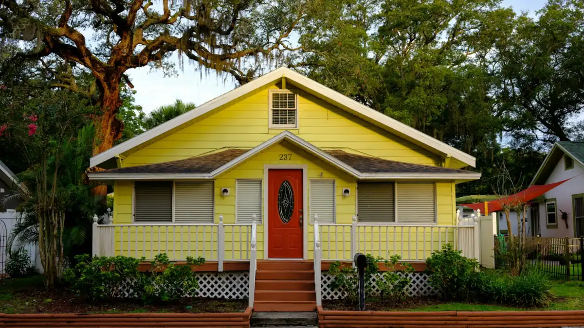 A yellow wooden house with a red door and a porch under live oak trees