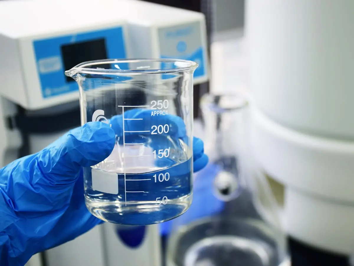 A gloved hand holding a graduated glass beaker of clear water in a laboratory